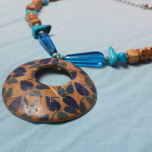 Ce collier bleu et marron possède un pendentif centrale en bois avec des motifs de feuilles dessus. Autour de celui ci est mis deux perles longues bleues en verre. Jusqu'à la fin du collier il y a ensuite une alternance entre de spetites perles bleues en turquoises, des perles bleues en nacres et des perles en bois carrées.