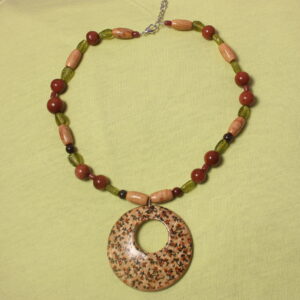 Ce collier est constitué d'un gros pendentif central rond. Sur celui ci se trouvent des petits détails verts et rouges en forme de fleur et de feuille. Autour de ce pendentif se trouve une alternance entre des perles de bois allongées, des perles marrons petites et d'autres grandes avec un verni pailleté sur celui ci qui ressort au soleil. et des perles de verre vertes en forme de feuilles.