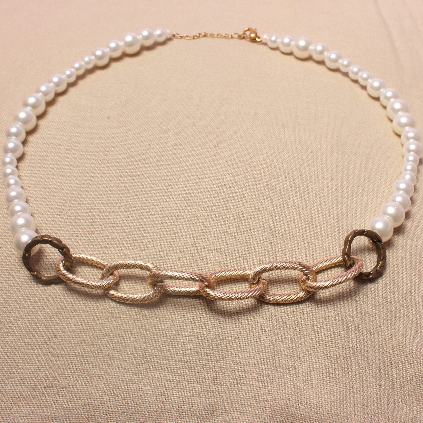 Ce collier maximaliste possède une grosse chaîne dorée en son centre. À l’extérieur de celle-ci se trouve une alternance de perles en verre ciré et de perles nacrées blanches. Les deux chaînettes sont séparées par un anneau doré foncé. Le collier se ferme grâce à un fermoir doré situé à son extrémité.