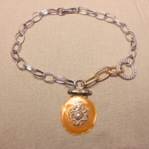 Ce collier est constitué de grosses chaînes autour d’un gros pendentif orange et rond. Celui-ci possède des détails en son centre. La chaîne est entièrement argentée du côté gauche, tandis qu’elle est asymétrique du côté droit. Elle possède un anneau argenté ainsi qu’une partie de chaîne dorée.