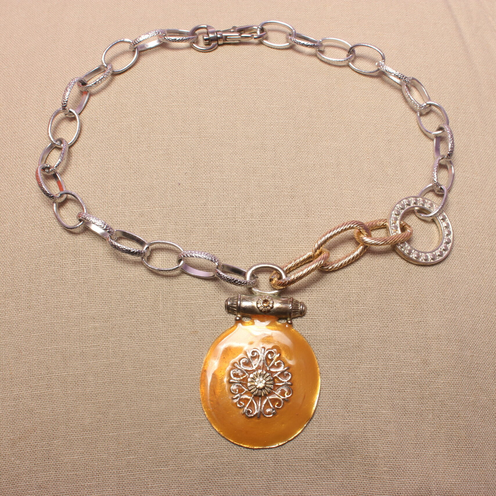 Ce collier est constitué de grosses chaînes autour d’un gros pendentif orange et rond. Celui-ci possède des détails en son centre. La chaîne est entièrement argentée du côté gauche, tandis qu’elle est asymétrique du côté droit. Elle possède un anneau argenté ainsi qu’une partie de chaîne dorée.