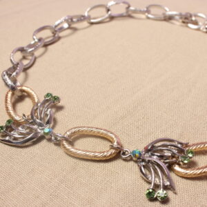 Ce gros collier est constitué de deux pendentifs en forme d’ailes de papillon, ornés de petites strass vertes. Entre ces deux ailes se trouve un anneau doré, qui se retrouve également de l’autre côté des ailes. Une grosse chaîne argentée se poursuit ensuite jusqu’à l’extrémité du collier. Celui-ci se ferme grâce à un fermoir situé au bout de la chaîne.