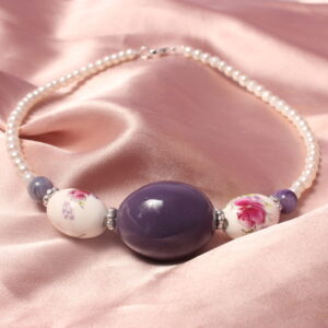 Ce collier est fait symétriquement autour d'une grosse perle violette en céramique. Autour de celle ci est mis deux perles ovales en céramique égalment. Ces deux perles possèdent des détails fleuris plutot détaillés. ces perles sont séparées par des petites intercalaires. Il y a ensuite une petite perle violette qui est suivie par des petites perles en verre cirées nacrées blanches qui se continuentjusqu'a la fin de collier. il se ferme grace a un petit fermoir et un anneau situé au bout du collier.