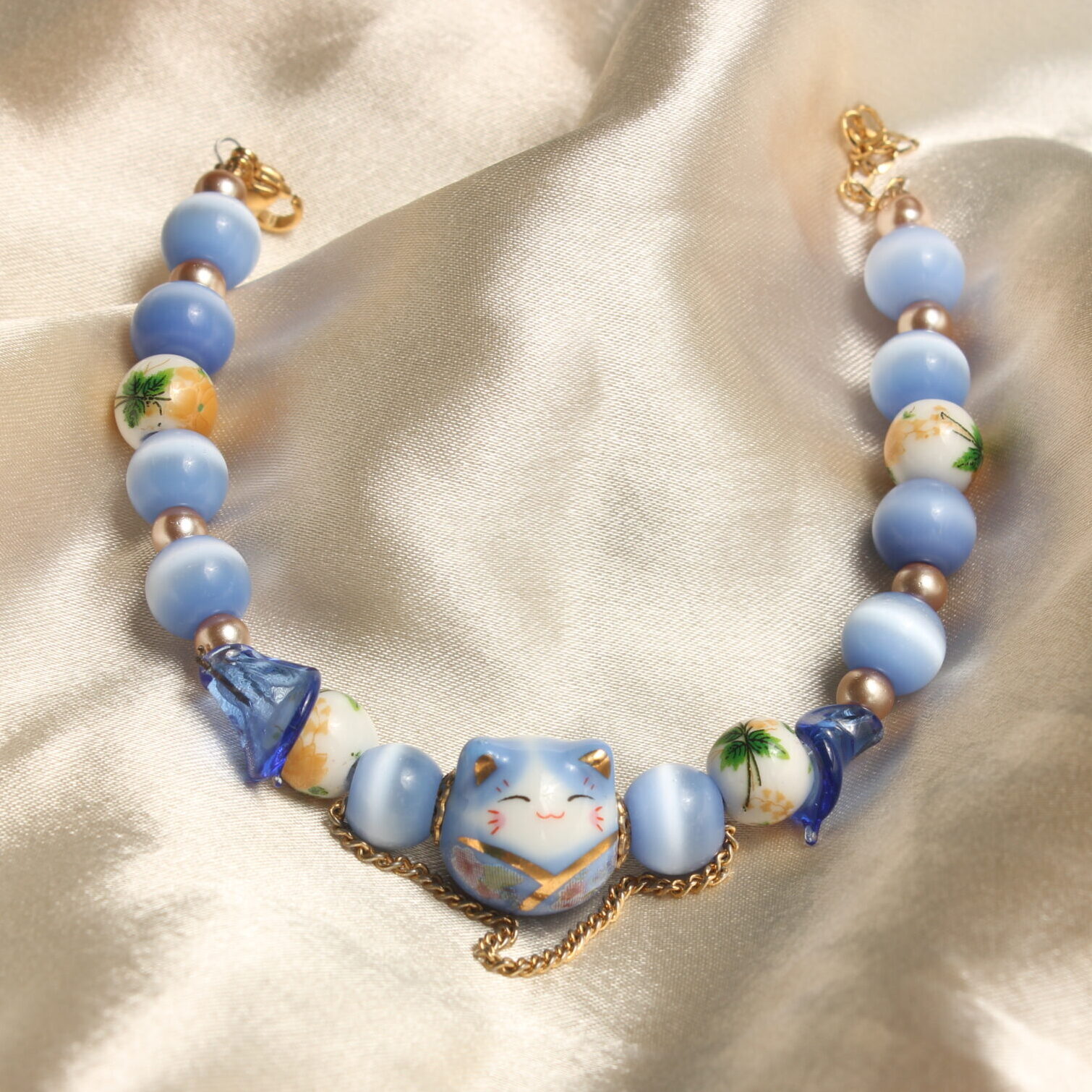 Ce bracelet est constitué d'une perle centrale en forme de chat bleu. Autour de celle ci est installée une succession de perles en verre œil de chat bleues claires, des perles de verres bleues en forme de fleur, des perles de céramique avec des détails fleuris dessus et des perles dorées en verre ciré. Ce bracelet se ferme grâce à un fermoir et une chaînette. Il y a également une petite chaînette décorative autour de la perle en forme de chat.