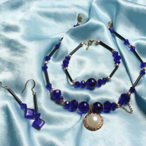 Cette parure de bijoux est constituée d'un bracelet, d'un collier et de boucles d'oreilles. Les boucles d'oreilles possèdent une perle en verre noire en forme de tube, suivie de perles en verre carrées bleues nuit. Le bracelet et le collier possèdent une alternance de perles en verre bleues nuit rondes, plates et losange. Ainsi que des perles dorées en verre ciré, en plus de perles en verre en forme de tube noires. Le collier possède également des petites chaînettes qui pendent légèrement et un pendentif central doré et blanc.
