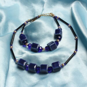 Cette parure de bijoux est constitué d'un bracelet et d'un collier. Ils contiennent tous les deux une succession de perles de céramique carrés bleues nuit. les perles plates en verre bleu nuit, des perles dorées en verre cirées et de perles noirs en tube.