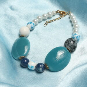 Ce bracelet est constitué d'une perle centrale bleue et blanche en céramique représentant des petites fleurs. autour ce celle ci sont mises des perles en pierre bleues foncées. Puis, deux intercalaires dorées sont installées autour de deux grosses perles bleues et ovales probablement en pierre. il y a ensuite une perle grise à droite ainsi qu'une perle blanche à gauche faisant une petite asymétrie au bracelet. Des perles en céramique sont installées avant de finir le bracelet avec des perles blanches et nacrées en perles de verre ciré.