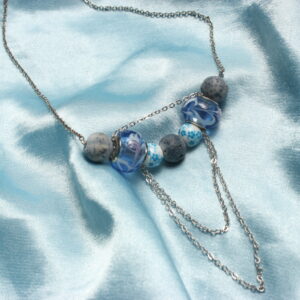 Ce collier est constitué de deux perles en verre détaillées de petits dessins fleuris. Autour de celles ci sont installées des perles de lave bleues ainsi que des perles en céramique avec des motifs floraux. Des petites chaînettes argentées pendent légèrement.