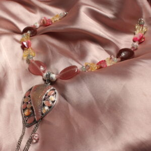 Ce collier est fabriqué symétriquement autour du gros pendentif central en forme de gouttelette d'eau rose. Celui ci possède des détails sur les cotés en forme ovale ou circulaire. Dessous celui ci sont accrochées des petites chaînettes ainsi que des petites perles. Autour de ce pendentif, il y a donc une suite de perles. Une première perle en céramique avec une fleur rose représentée dessus. puis deux petites intercalaires en forme de fleur sont installées. il y a après une grosse perle rose en verre de forme ovale. Les deux intercalaires reviennent ici. puis, une perle nacrée rose en forme de losange est entourée par deux perles jaunes en forme de papillon ainsi que de deux perles bleu/ grise en verre. ici il y a une petite chaînette disposée sur le coté droit qui tombe et du coté gauche sont mises des petites perles en verre en forme de gouttes d'eau. ensuite, deux intercalaires entourent une grosse perle marron ovale en verre ou en pierre. Les intercalaires roses continuent d'entourer chaque perles jusqu'à la fin du collier. Il y a donc une petite perle en céramique avec une fleur représentée dessus, puis une perle losange nacrée rose, une perle en verre jaune papillon et enfin une petite perle bleu/ grise. Ce collier est alors fermé avec un petit fermoir et une chaînette avec un petit papillon au bout de celui ci.