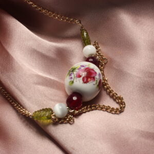 Ce joli collier est construit de manière symétrique autour d’une grosse perle centrale ronde en céramique. Celle-ci est blanche et ornée de motifs floraux détaillés. Autour de celle-ci sont disposées une petite perle violette, probablement en pierre, une autre perle blanche, également probablement en pierre, ainsi qu’une perle en verre verte en forme de feuille. Le collier se termine par une chaîne et se ferme grâce à un fermoir. Il possède également de petites chaînettes disposées au centre, qui tombent légèrement.