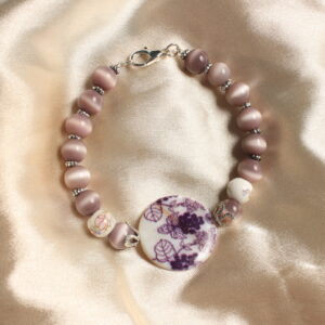 Ce bracelet est constitué d'une perle centrale en céramique violette. Celle ci possède des détails avec des motifs représentant des grappes de raisin. autour de celle ci se trouvent des intercalaires argentés. Il y a ensuite une succession de perles de verre œil de chat violettes. Elles sont toutes séparées de petites intercalaires. Il y a également deux perles de céramique avec des petits détails fleuris dessus. Fermoir argenté