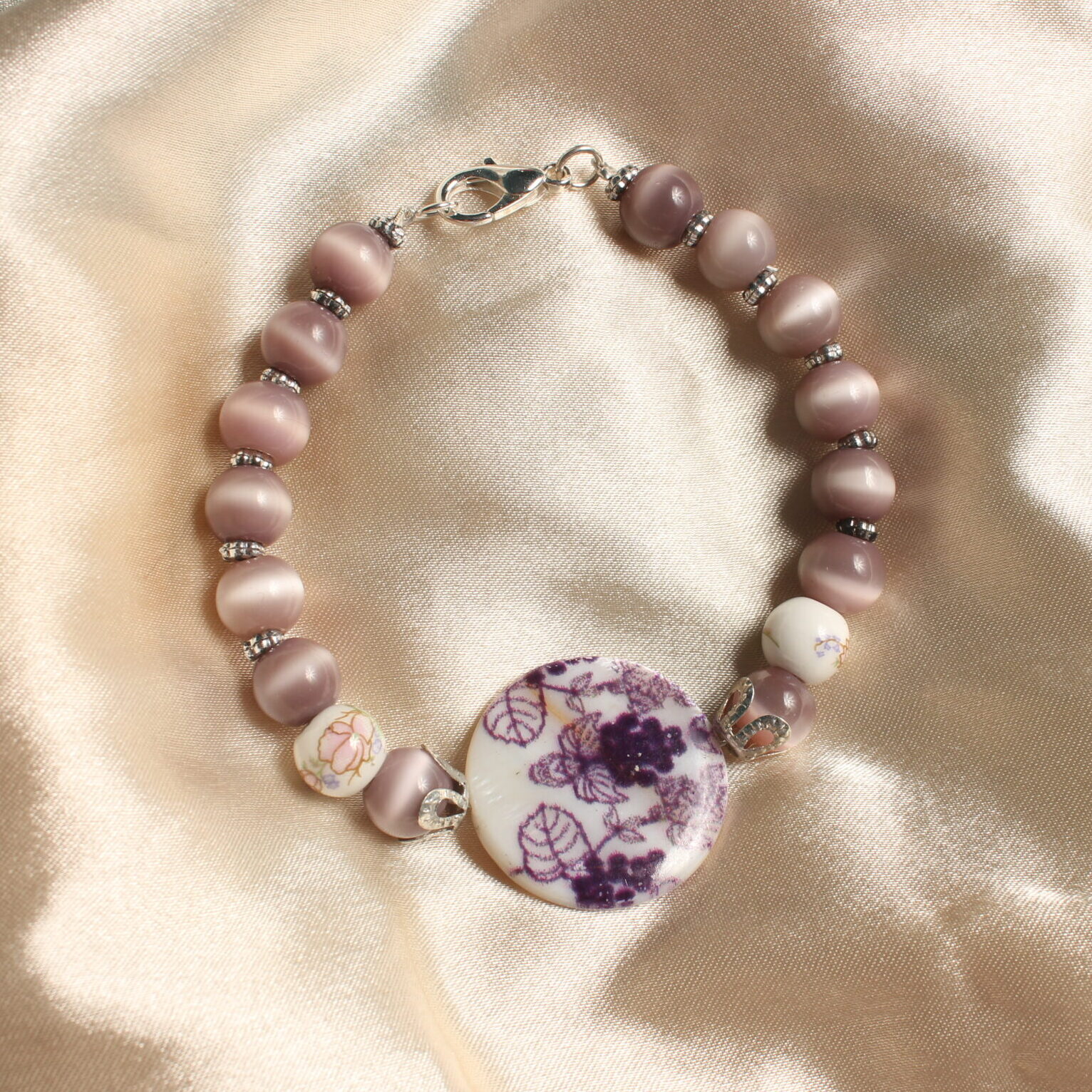 Ce bracelet est constitué d'une perle centrale en céramique violette. Celle ci possède des détails avec des motifs représentant des grappes de raisin. autour de celle ci se trouvent des intercalaires argentés. Il y a ensuite une succession de perles de verre œil de chat violettes. Elles sont toutes séparées de petites intercalaires. Il y a également deux perles de céramique avec des petits détails fleuris dessus. Fermoir argenté