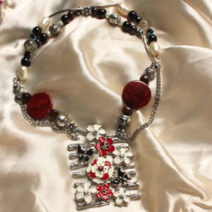 Ce collier est disposé symétriquement autour d'un gros pendentif central. Celui ci possède 7 barres verticales avec des petits détails imitant le bois. sur celui ci sont représentés des petites fleurs et des petits papillons rouges, blancs et noirs. Le pendentif est posé sur une longue perle argentée. Ensuite, une grosse perle rouge et aplatie est entourée d'une petite perle en pierre marron, d'une grosse perle argentée et d'une petite perle en forme de fleur. ceci est également entouré par une petite chaînette. Puis, il y a encore une longue perle argentée. Puis, une perle argentée est ensuite entourée par une petite perle nacrée blanche' une perle noire, une perle en pierre marron, et d'une perle en forme de fleur. Il y a ensuite une autre longue perle argentée. Enfin, on a une perle grise qui est entourée de deux petites perles irisées noires, d'une perle noire et d'une perle ovale nacrée blanche. Celui-ci est fermé grâce à un fermoir et un anneau.