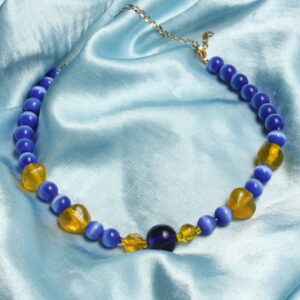 Ce collier possède une grosse perle centrale en verre bleue nuit. Autour de celle ci se trouve une succession de perle en verre jaune de différentes forme ainsi que des perles œil de chat. Ce collier se ferme grâce à un fermoir et une chaînette dorée.