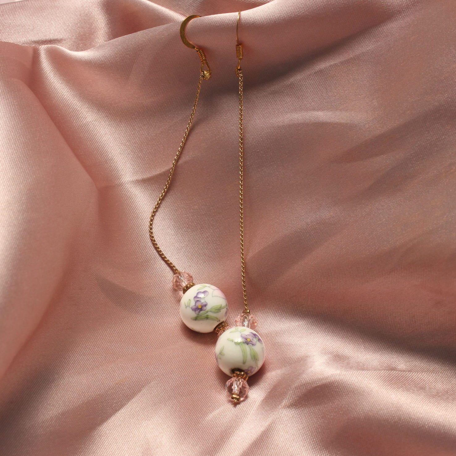Ces boucles d'oreilles sont constituées d'un fil doré ainsi que trois perles. deux roses en verres autour d'une plus grosse en céramique avec des détails fleuris roses verts et violets.