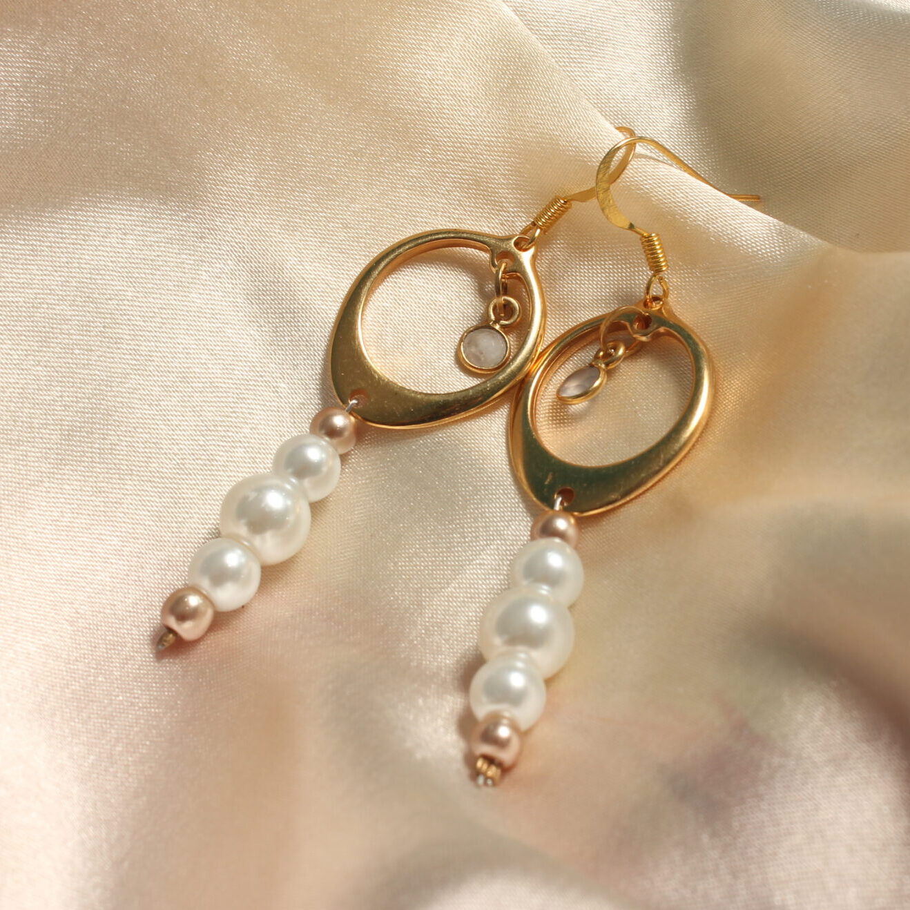Ces boucles d’oreilles dorées sont constituées d'un anneau doré ainsi qu'in petit pendentif gris en son centre. Des perles nacrées en verres cirées de différentes tailles blanches et dorées pendent légèrement.